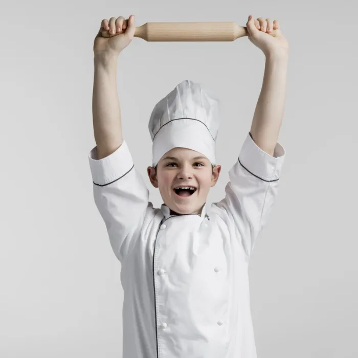 A smiling child in a chef’s hat holding a rolling pin above their head
